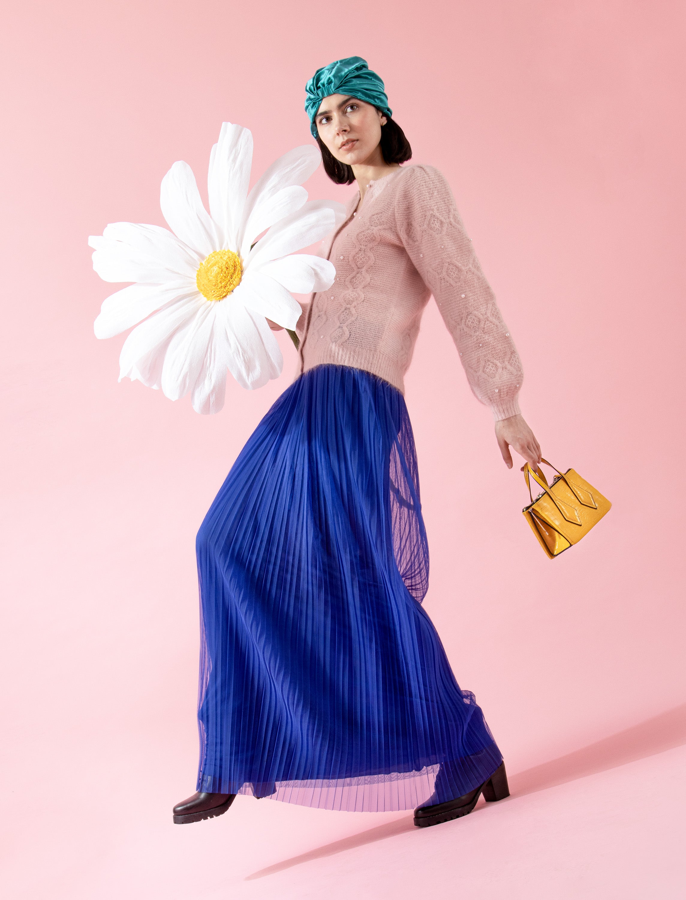 Woman in a blue pleated skirt and beige top holding a large white flower against a pink background